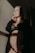 A pregnant woman stands in soft lighting, wearing a black lace outfit and a dark robe. Her hand rests under her belly, and she gazes thoughtfully into the distance. The light casts a warm, gentle glow on her face and the wall behind.