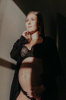 A pregnant woman stands in soft lighting, wearing a black lace outfit and a dark robe. Her hand rests under her belly, and she gazes thoughtfully into the distance. The light casts a warm, gentle glow on her face and the wall behind.