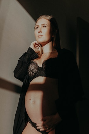 A pregnant woman stands in soft lighting, wearing a black lace outfit and a dark robe. Her hand rests under her belly, and she gazes thoughtfully into the distance. The light casts a warm, gentle glow on her face and the wall behind.
