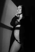 A pregnant woman stands partially in shadow, wearing a lace bra and draped in an open robe. She gazes upward with a serene expression, one hand gently resting on her belly. The lighting creates dramatic contrasts with sharp lines of light and shadow.
