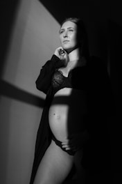 A pregnant woman stands partially in shadow, wearing a lace bra and draped in an open robe. She gazes upward with a serene expression, one hand gently resting on her belly. The lighting creates dramatic contrasts with sharp lines of light and shadow.