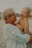 An older woman with glasses is holding a young child who is laughing joyously. They are both dressed in light clothing, suitable for warm weather or a beach setting. The background is out of focus, giving a serene and casual atmosphere.