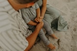 A child and an adult are sitting on the sand. The child is wearing a grey outfit and the adult is wearing a striped shirt. The adult is holding the child's hands, both sitting closely together with visible affection.