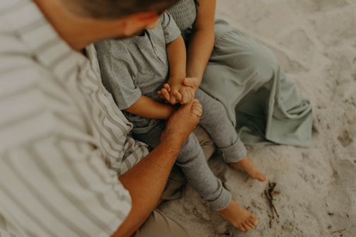 A child and an adult are sitting on the sand. The child is wearing a grey outfit and the adult is wearing a striped shirt. The adult is holding the child's hands, both sitting closely together with visible affection.