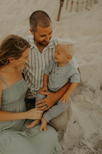 A family consisting of a man, woman, and a young child sits on a sandy beach. The man wears a striped shirt and holds the child on his lap, who is dressed in a gray outfit. The woman, wearing a light green dress, is seated beside them, tenderly engaging with the child. Their expressions reflect warmth and connection.