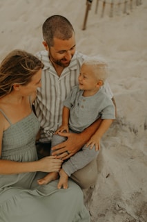 A family consisting of a man, woman, and a young child sits on a sandy beach. The man wears a striped shirt and holds the child on his lap, who is dressed in a gray outfit. The woman, wearing a light green dress, is seated beside them, tenderly engaging with the child. Their expressions reflect warmth and connection.