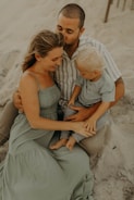 A family sitting together on a sandy beach, with an adult man embracing a woman and child. The woman is wearing a long, flowing light green dress, while the child is dressed in a gray outfit. The setting seems peaceful and intimate, with a warm and relaxed atmosphere.