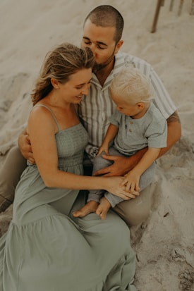 A family sitting together on a sandy beach, with an adult man embracing a woman and child. The woman is wearing a long, flowing light green dress, while the child is dressed in a gray outfit. The setting seems peaceful and intimate, with a warm and relaxed atmosphere.