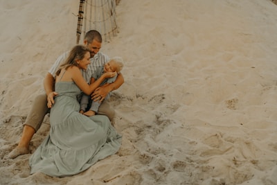 A family is sitting together on a sandy beach. The group includes an adult male, an adult female, and a young child. They are sitting closely, with the female embracing the child, who is smiling. The background features sand dunes and a part of a wooden fence.