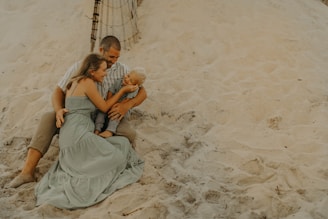 A family is sitting together on a sandy beach. The group includes an adult male, an adult female, and a young child. They are sitting closely, with the female embracing the child, who is smiling. The background features sand dunes and a part of a wooden fence.