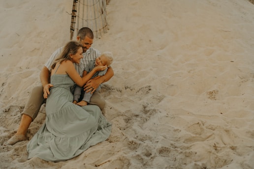 A family is sitting together on a sandy beach. The group includes an adult male, an adult female, and a young child. They are sitting closely, with the female embracing the child, who is smiling. The background features sand dunes and a part of a wooden fence.