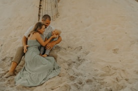 A family is sitting together on a sandy beach. The group includes an adult male, an adult female, and a young child. They are sitting closely, with the female embracing the child, who is smiling. The background features sand dunes and a part of a wooden fence.