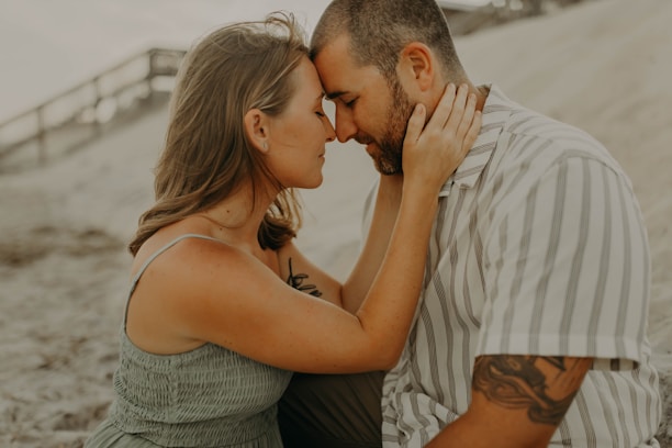 A couple sits closely together on a sandy beach, with the woman gently placing her hands on the man's face. They are touching foreheads, creating an intimate and tender moment. The background features a blurred view of a boardwalk and soft sand dunes.