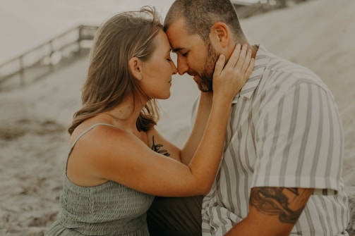 A couple sits closely together on a sandy beach, with the woman gently placing her hands on the man's face. They are touching foreheads, creating an intimate and tender moment. The background features a blurred view of a boardwalk and soft sand dunes.