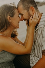 A couple is sitting closely together, with their foreheads touching and smiling warmly at each other. The woman is gently holding the man's face with both hands. The scene conveys a sense of affection and intimacy.