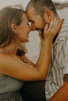 A couple is sitting closely together, with their foreheads touching and smiling warmly at each other. The woman is gently holding the man's face with both hands. The scene conveys a sense of affection and intimacy.