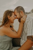 A couple is seated closely together, gently touching foreheads and smiling warmly at each other. The man has a trimmed beard and is wearing a striped shirt, while the woman has long hair and is wearing a sleeveless dress.