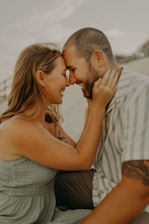 A couple is seated closely together, gently touching foreheads and smiling warmly at each other. The man has a trimmed beard and is wearing a striped shirt, while the woman has long hair and is wearing a sleeveless dress.