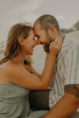 A couple is seated closely together, gently touching foreheads and smiling warmly at each other. The man has a trimmed beard and is wearing a striped shirt, while the woman has long hair and is wearing a sleeveless dress.