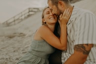 A couple is sitting on the sand, sharing a joyful and intimate moment together. The woman is smiling broadly with her eyes closed, while affectionately holding the man's face. The man is leaning in close to her, with a gentle expression. The setting appears to be a beach, with a blurred background of sand and a wooden pathway.