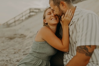 A couple is sitting on the sand, sharing a joyful and intimate moment together. The woman is smiling broadly with her eyes closed, while affectionately holding the man's face. The man is leaning in close to her, with a gentle expression. The setting appears to be a beach, with a blurred background of sand and a wooden pathway.