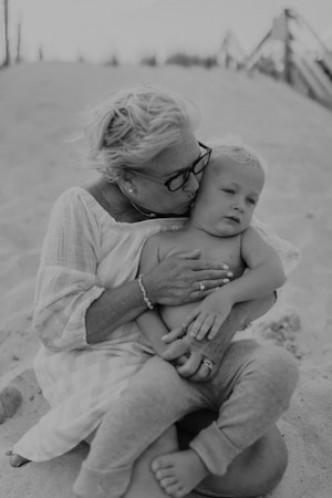 A woman with short hair and glasses embraces a young child while sitting on a sandy beach. The child looks relaxed, and the woman affectionately kisses the child's forehead. Both are dressed in casual, light-colored clothing.