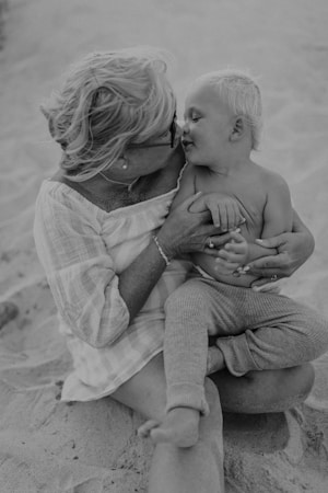 A black-and-white photograph depicts an older woman hugging a toddler. They are sitting on sandy ground, the woman has short hair and is wearing glasses and a light, long-sleeved top. The toddler appears to be relaxed and is only wearing pants. They are gazing at each other lovingly.