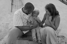 A family enjoying time together on a sandy beach. The group consists of two adults, sitting on either side of a young child. All three are smiling and appear relaxed, suggesting a moment of joy and connection. The adults seem to be wearing casual, comfortable clothing.