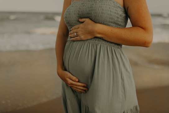 A person in a light, flowing dress cradles their belly tenderly, with one hand resting above and the other below, standing on a beach with gentle waves in the background.