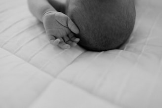 A black and white photograph captures the gentle, close-up image of an infant's hand and head resting on a soft, quilted surface. The delicate details of the baby's skin and hair create a sense of intimacy and tenderness.