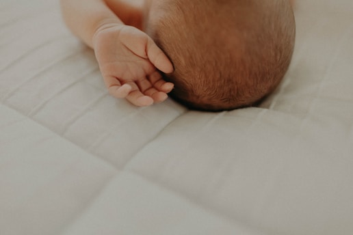 A close-up of a baby's head and hand resting on a soft, quilted surface. The baby's hand is relaxed and resting beside the head, revealing delicate fingers. The scene is warmly lit, highlighting the soft textures of the baby's skin and the quilt.