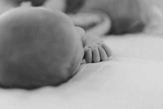 A black and white photograph of a baby lying down, focusing on their small hand with curled fingers. The soft focus adds a calming and serene quality to the image.