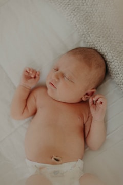 A sleeping newborn baby lies on a soft, textured blanket with a visible umbilical cord stump and wearing a diaper. The baby has a peaceful expression, slightly closed eyes, and small clenched fists.