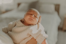 A newborn baby is peacefully sleeping, dressed in a cozy knitted sweater. The baby is being gently held, with a soft background of a bed and pillows blurred out, giving the scene a serene and warm feeling.