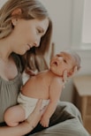 A woman gently cradles a newborn baby in her arms, looking at the infant with a serene and tender expression. The baby is wrapped in a diaper, and both are in a softly lit indoor setting.