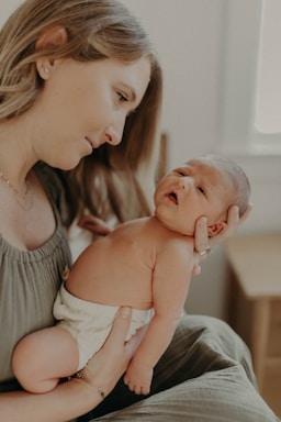 A woman gently cradles a newborn baby in her arms, looking at the infant with a serene and tender expression. The baby is wrapped in a diaper, and both are in a softly lit indoor setting.