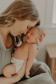 A tender moment between a woman and a newborn baby. The woman gently holds the sleeping baby against her shoulder, with her eyes closed and a serene expression. The setting appears soft and intimate, with light tones enhancing the warm atmosphere.