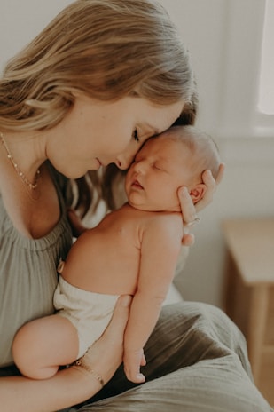A tender moment between a woman and a newborn baby. The woman gently holds the sleeping baby against her shoulder, with her eyes closed and a serene expression. The setting appears soft and intimate, with light tones enhancing the warm atmosphere.