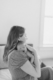A woman is holding a sleeping newborn baby close to her chest in a tender embrace. The setting appears calm and serene with soft lighting and gentle expressions.