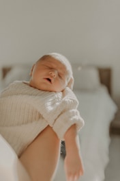 A peaceful, sleeping baby is gently cradled in someone's arms, wearing a soft, knitted cream sweater. The lighting is warm and soft, creating a serene and intimate atmosphere. In the background, a blurred bed adds a sense of calm domesticity.
