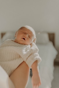 A peaceful, sleeping baby is gently cradled in someone's arms, wearing a soft, knitted cream sweater. The lighting is warm and soft, creating a serene and intimate atmosphere. In the background, a blurred bed adds a sense of calm domesticity.