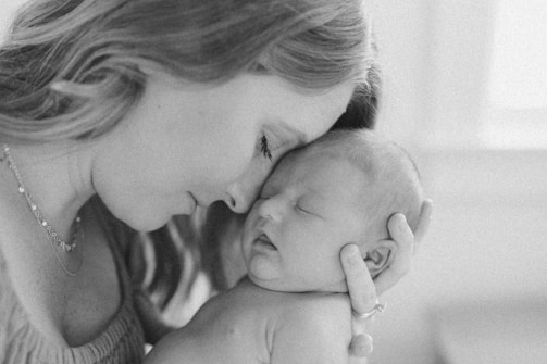 A tender black and white photograph of a mother holding her newborn baby gently against her face, eyes closed in a serene moment of intimacy.
