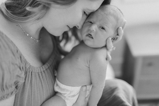 A tender moment between a woman and a newborn baby captured in close-up. The woman is gently holding the baby, who appears calm and content. Both are embraced in a warm and intimate pose, with soft focus on their faces, creating a loving atmosphere.