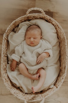 A baby is peacefully sleeping in a woven basket lined with soft, cream-colored padding. The baby is dressed in a cozy, knitted sweater and has a calm expression on its face.