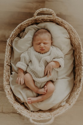 A newborn baby, peacefully asleep in a cozy, woven basket lined with soft, cream-colored fabric. The baby is dressed in a knitted cream sweater and appears comfortable and serene.