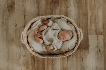 A sleeping baby dressed in a knitted outfit is peacefully nestled in a wicker basket lined with soft cushions. The basket is placed on a wooden floor, and the overhead view highlights the baby's serene posture and the cozy setting.