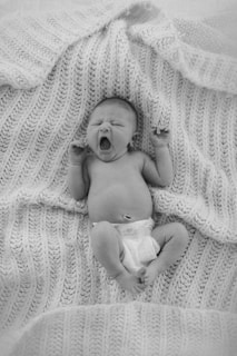 A newborn baby lies on a cozy, knitted blanket, yawning with arms raised. The baby is wearing a diaper and appears peaceful and relaxed.