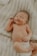 A newborn baby lies on a soft, knitted blanket. The baby has a peaceful expression with eyes closed and a slight smile. One hand is raised slightly while the other rests on the baby's tummy. The diaper is visible, and the background provides a cozy texture.