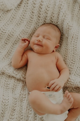 A newborn baby lies on a soft, knitted blanket. The baby has a peaceful expression with eyes closed and a slight smile. One hand is raised slightly while the other rests on the baby's tummy. The diaper is visible, and the background provides a cozy texture.
