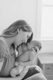 A woman with long hair gently holds a newborn baby in her arms, gazing lovingly at the infant. The baby, wearing a diaper, is cradled closely to her chest. Both the woman and the baby share a tender, intimate moment, creating a serene and heartwarming scene. The image is in black and white, adding a classic and timeless feel.